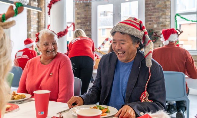Two Crisis members are sat eating Christmas lunch together. They are smiling and laughing. The room has Christmas decorations up and the members are wearing festive hats.