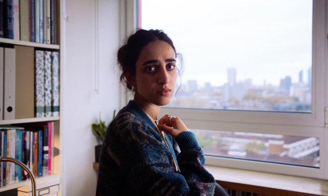 A  young woman standing by a window in a tower block of flats, looking directly at the camera.