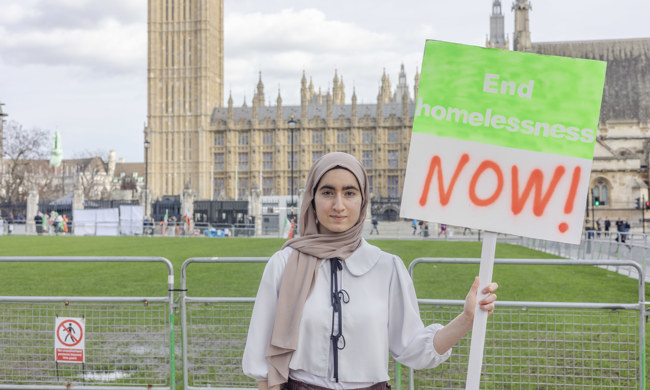 A woman wearing a headscarf stands outside Parliament holding a placard that says 'End homelessness NOW!'