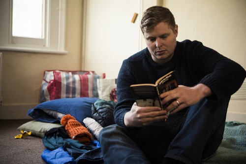 Man reading a book sitting on a makeshift bed on the floor