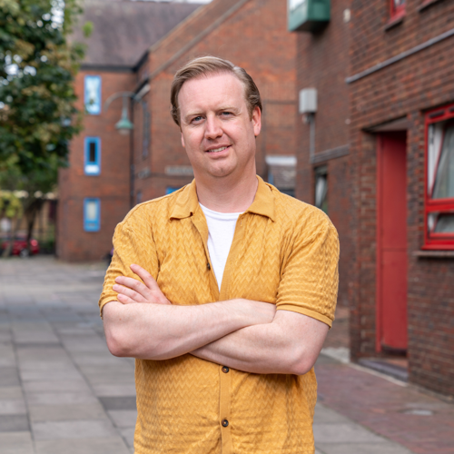 A man wearing a yellow shirt stands with his arms crossed in a street with brick houses and flats behind him