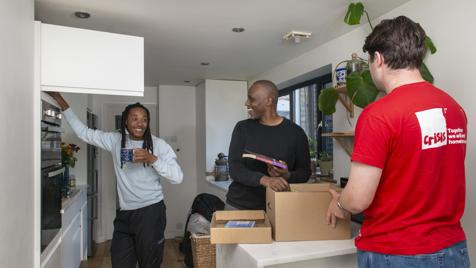 Three men are standing in a kitchen. They are unpacking boxes and chatting with one another.
