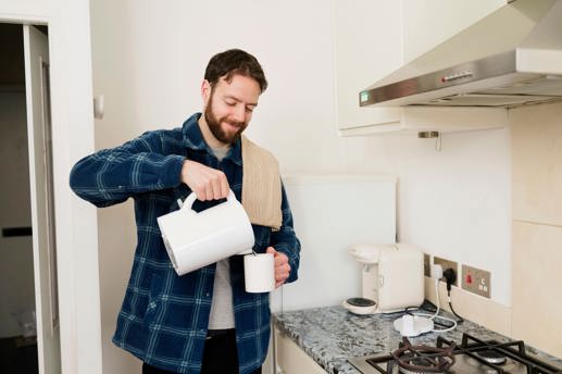 Bradley stands in a kitchen pouring from a kettle into a mug. He looks content.